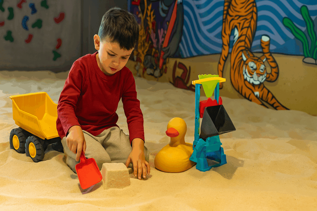 A child in a sandbox confidently selecting tools and building a simple sand structure in an indoor sandbox playground setting